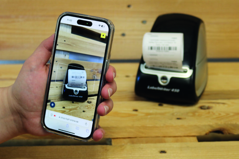 Hand mit Handy in der Hand, Etikettendrucker mit Etiketten, ScanApp auf dem Handy